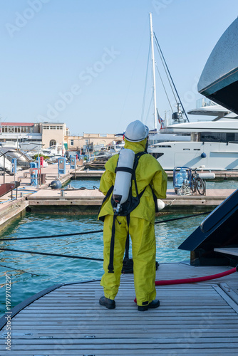 Fire drill on yacht stern: crew member in protective suit uses fire hose and breathing apparatus during training, simulating emergency firefighting on board.