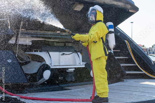 Fire drill on yacht stern: crew member in protective suit uses fire hose and breathing apparatus during training, simulating emergency firefighting on board.