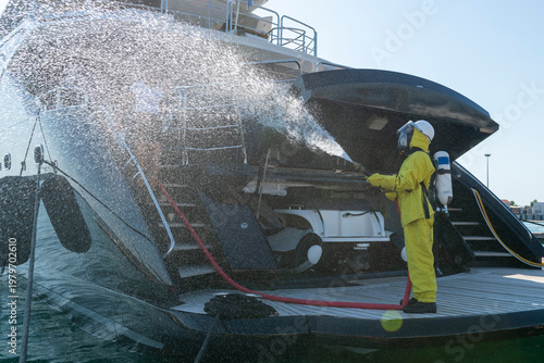 Fire drill on yacht stern: crew member in protective suit uses fire hose and breathing apparatus during training, simulating emergency firefighting on board.