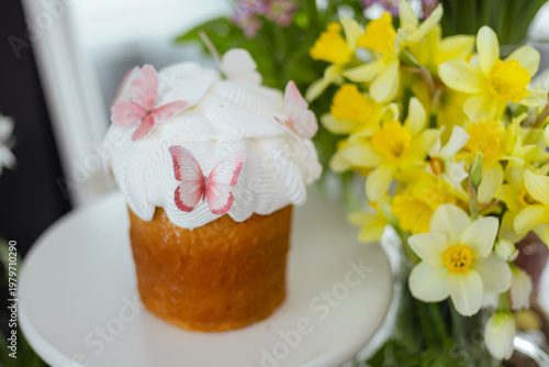 Easter cake with white icing and butterfly decoration surrounded by spring flowers, bright festive composition with copy space on light background.