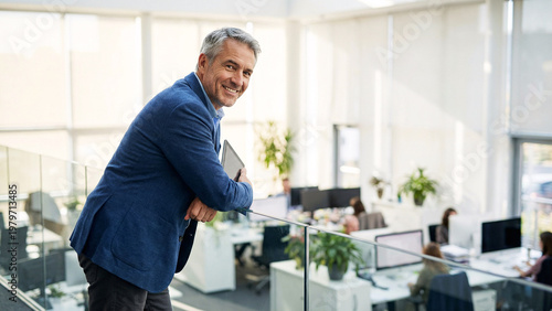 Experienced Male Advisor Leaning Against Hallway Railing In Contemporary Office Atrium, Tablet Under Arm, Blurred Workstations, Diffused Morning Light, Executive Workplace