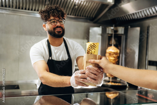 Male food vendor in black apron hands over wrapped kebab to customer at counter in modern fast food restaurant with grill in the background