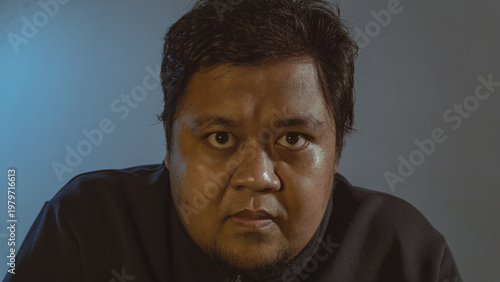 An intense adult male wearing a dark casual jacket stares directly into the camera with a highly serious and unwavering facial expression. The dramatic cinematic lighting strongly