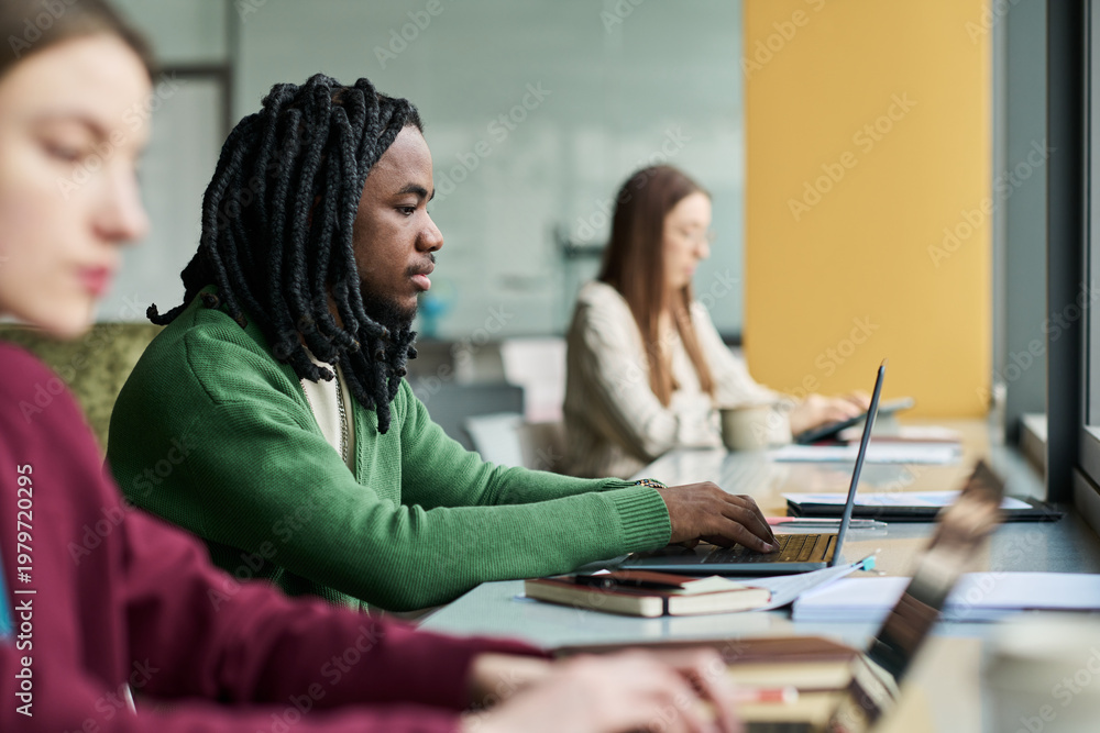 Fototapeta premium Young African American man working on laptop in shared office with colleagues, supporting teamwork and productivity. Ideal for business training, coworking, recruitment, or workplace communication