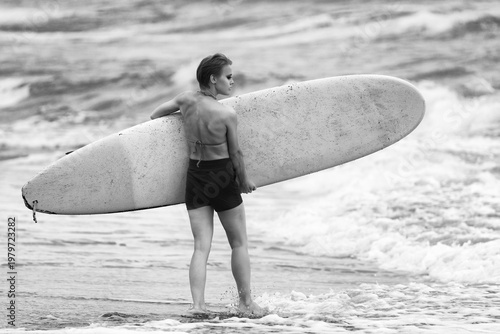 фотографія Black and white rear view of strong athletic woman with short hair holding surfboard
