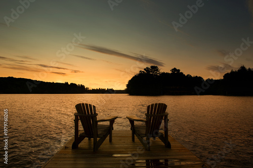 Two Adirondack chairs on wooden dock overlooking calm lake at golden sunset with silhouetted pine trees and warm orange sky reflections on water in Muskoka Ontario cottage country summer evening