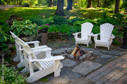 White Adirondack chairs arranged around rustic stone fire pit on flagstone patio surrounded by lush green hostas in shaded woodland garden setting during warm summer afternoon in Muskoka Canada