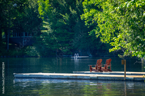 Two red Adirondack chairs on wooden dock overlooking calm lake with lush green trees in warm summer sunlight, serene cottage country retreat in Muskoka Ontario Canada with peaceful water reflections