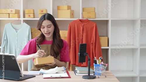 A small startup business run by a young Asian woman working from home, using a laptop for online marketing and preparing packaging for customer orders.