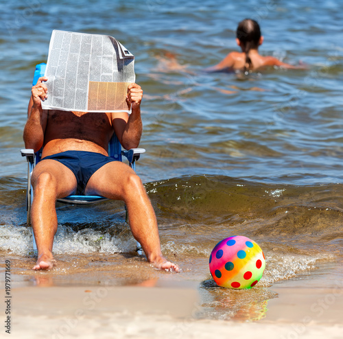 A man is relaxing in an armchair right by the waves, engrossed in his reading and covering his face with an open newspaper