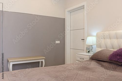 A minimalist bedroom with a beige quilted bed, white tufted headboard, nightstand with a pleated lamp, a light beige bench, and a neutral-toned wall. Soft, calming palette.