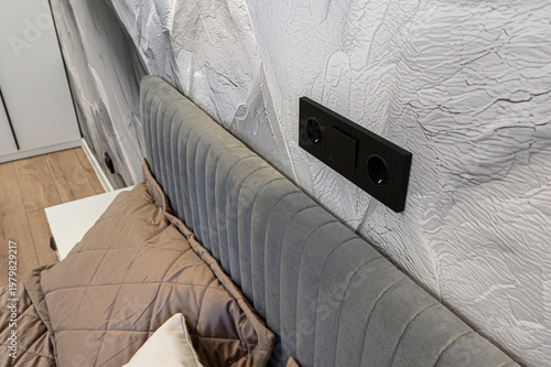 A modern bedroom corner with a grey upholstered headboard, textured grey wall, black wall sockets, beige quilted bedding, and a glimpse of a white nightstand on wooden flooring.