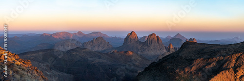 Elevated sunset view from Assekrem summit of the Atakor mountain chain. Jagged volcanic peaks glow orange against a soft gradient colorful sky in the Hoggar plateau of Tamanrasset, Algeria.