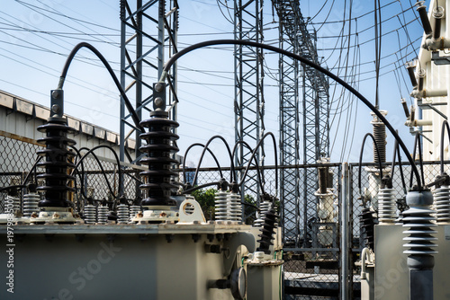 Large electrical substation with high-voltage equipment under bright blue sky. Black insulators and curved conductors dominate the scene. Metal towers stretch upward, carrying power lines