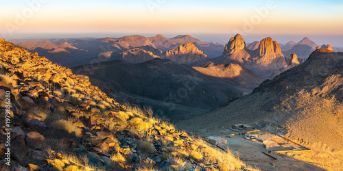 Elevated sunset view from Assekrem summit of the Atakor mountain chain. Jagged volcanic peaks glow orange against a soft gradient colorful sky in the Hoggar plateau of Tamanrasset, Algeria.