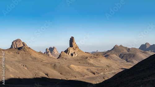 Elevated panorama of the Atakor chain in the Hoggar region. A sharp granite spire rises over the terrain with a dusty road leading toward Assekrem under a clear blue sky.
