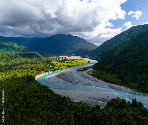 Aerial view of the Hokitika River carves through a landscape of dense, emerald forests meeting rugged mountains under a dynamic sky, Hokitika Gorge, West Coast Region, New Zealand.