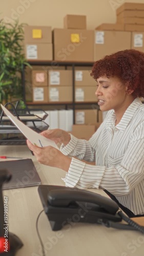 Woman reading a document and holding paper while touching her forehead amid stacked cardboard boxes and parcels at an office building, phone on desk; stressed focus.