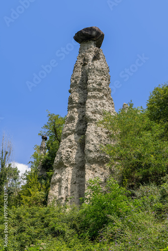 Monumental hoodoo at Pyramids of Zone near Lake Iseo under clear summer sky, pale sediment tower dominates green hillside while smaller capped spire behind it adds depth