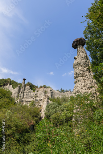 Wide vertical scene of Pyramids of Zone near Lake Iseo with dominant white earth tower, smaller hoodoos and chalky walls recede through dense greenery while dark capstone stands