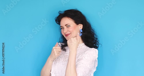 Cheerful woman waving hand and making inviting gesture against blue background, happy girl in white lace blouse smiling and welcoming, friendly studio portrait, communication concept.