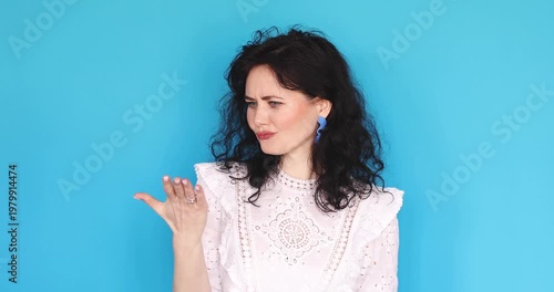 Young curly woman showing money gesture with fingers, asking for payment or cash, looking skeptical and demanding with expressive face on blue background, brunette lady making pay me sign