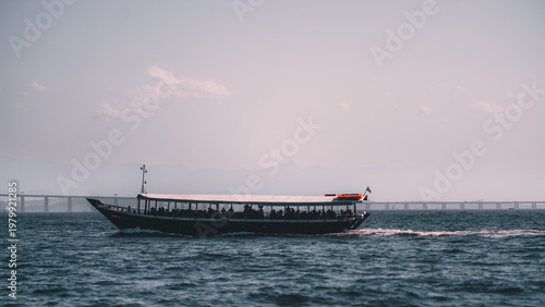 Telephoto view of passenger or tourist boat crossing open water at sunset, with dark silhouette figures onboard, gentle wake, distant bridge, hazy mountain backdrop and pastel sky with copy space