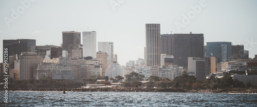 Telephoto view of modern urban skyline across calm waterfront, with dense high-rise office towers, hazy daylight, coastal cityscape, layered architecture and distant shore