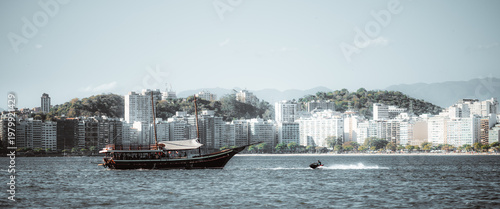Telephoto coastal cityscape with traditional schooner and speeding personal watercraft on open bay, framed by dense waterfront skyline, wooded hills and hazy mountain backdrop
