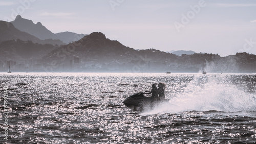 Silhouette riders on personal watercraft racing across sparkling bay, with dramatic spray, hazy mountain coastline, distant shoreline and bright backlit water at dusk