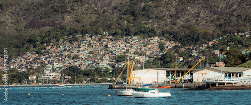 Telephoto view of coastal hillside community with dense favela housing, marina buildings, anchored sailboats and rocky tropical slope, capturing urban shoreline and layered neighborhood texture