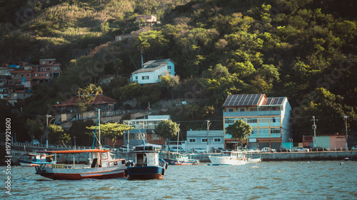 Telephoto view of small fishing boats on coastal water below a lush hillside village, with scattered houses, sunlit greenery, waterfront road and layered tropical landscape