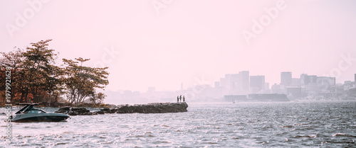 Telephoto coastal scene with yacht near rocky breakwater, dark silhouette anglers on the jetty, hazy city skyline across the bay, pastel sky and shimmering water