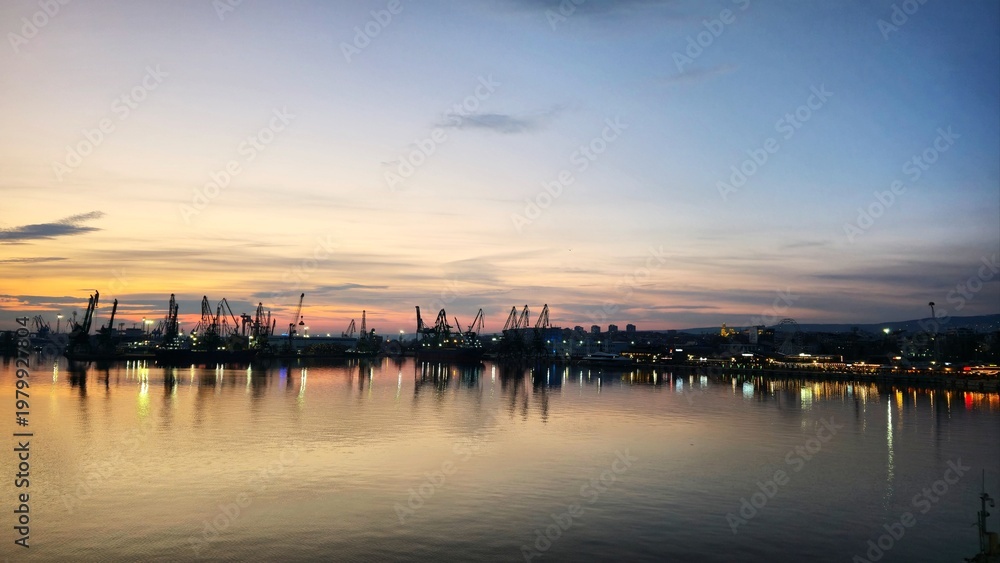 Fototapeta premium evening harbor panorama with glowing lights and cranes mirrored in water creating atmospheric urban waterfront scene with soft pastel sky tones