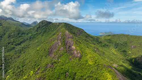 A verdant mountain peak covered with dense vegetation, rising sharply under a clear blue sky with some clouds. Seychelles, Mahe.