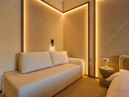 Modern hotel room corner featuring a beige sofa, bed, and wall panels with warm LED lighting. Minimalist interior design with wooden accents and cozy atmosphere.