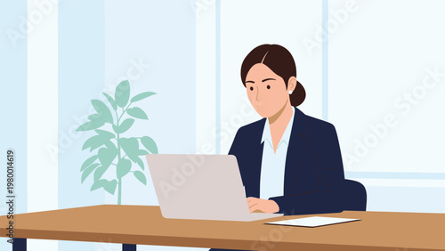 Professional woman with dark hair tied back wearing a dark blue suit jacket working on her laptop at a wooden desk in an office.