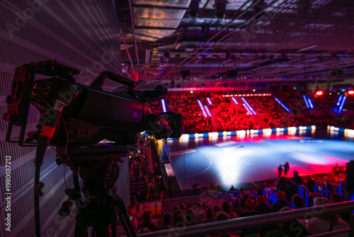Fotografie Professional broadcast camera on a tripod set up in a large sports arena, focused on a dimly lit handball court with spectators