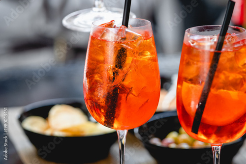 Vibrant orange Spritz cocktails with ice and citrus slices served in wine glasses with straws, accompanied by snacks in a casual dining setting