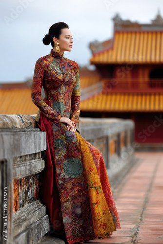 Asian woman in traditional Vietnamese ao dai dress with intricate patterns stands gracefully on a stone wall, historic architecture visible in the background