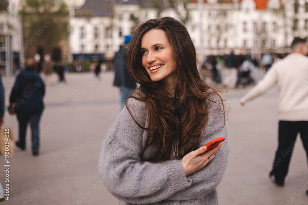 Fototapeta premium Playful young woman with long brunette hair looking away and smiling while holding red smartphone on city square, happy girl in grey sweater enjoying urban atmosphere and digital lifestyle