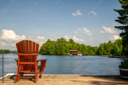 A brown Adirondack chair sits on a weathered wooden dock facing a calm blue lake, a pine-covered island with a cottage visible across the water on a sunny summer day in Ontario, Canada.