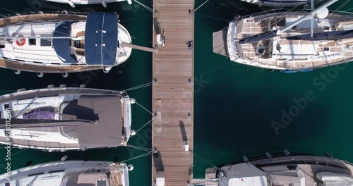 Overhead View Of Yachts Moored Along Pier With Lone Dockhand Walking, Turquoise Water, Calm Reflections And Orderly Berths Creating Geometric Pattern