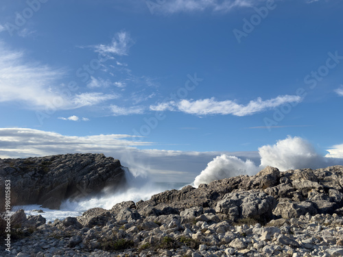 Atlantic wave energy creating sequential impact along rocky coast. Foam disperses into fine spray under deep blue sky with natural copy space for banner background