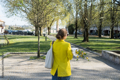 Fotografie Rear view of a woman in a yellow coat strolling through a sunny park area, holding a bunch of lemons, symbolizing freshness and spring