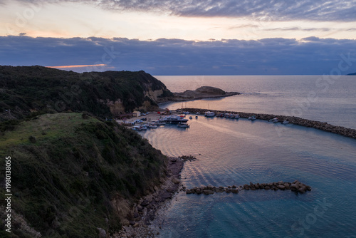 Aerial view of the small fishing harbor of Agios Stefanos Avliotes at sunset, featuring stone breakwaters, moored boats, and calm turquoise waters on the northwest coast of Corfu island, Greece.