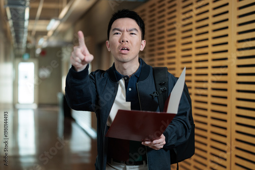 Quadro su tela A lost newcomer Asian male student with a backpack holds a folder while pointing and expressing frustration in a university hallway, ideal for international student, orientation, freshman concepts