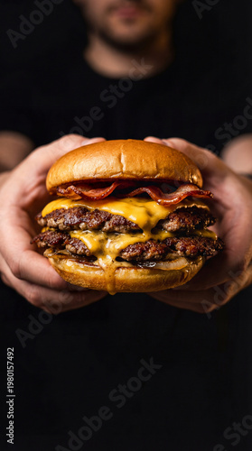 Man holding a loaded bacon cheeseburger against a dark background. Moody food photography with melted cheese and crispy bacon.