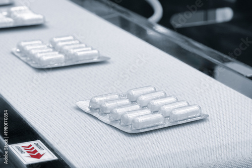 Blister packs move along a factory conveyor belt. White tablets sit neatly in plastic packaging. Industrial setting shows pharmaceutical production. Image fits medical