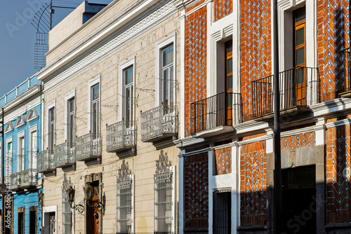 Historic street lined with colorful colonial facades featuring ornate wrought iron balconies and traditional Talavera tiles under bright blue sky in Puebla, Mexico